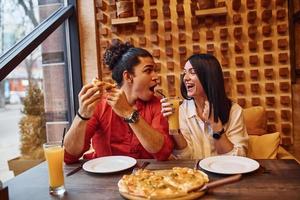 multiethnisches junges paar sitzt drinnen zusammen und isst pizza foto