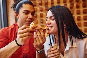 multiethnisches junges paar sitzt drinnen zusammen und isst pizza foto