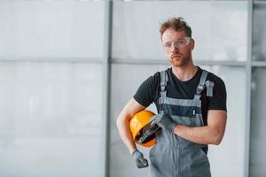 Porträt eines Mannes in grauer Uniform und mit orangefarbenem Schutzhelm in den Händen, der tagsüber in einem modernen großen Büro drinnen steht foto