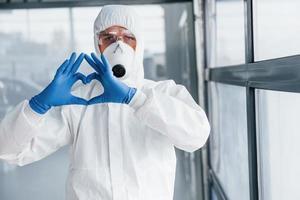männlicher arztwissenschaftler in laborkittel, defensiver brille und maske zeigt liebesgeste mit den händen foto