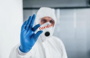 männlicher arztwissenschaftler in laborkittel, defensiver brille und maske hält glas mit dem covid-19-wort darauf foto