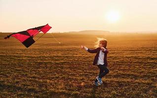 glückliches kleines Mädchen, das zur Sonnenaufgangszeit mit Drachen in den Händen auf dem schönen Feld läuft foto