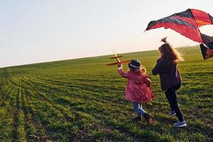 Zwei kleine Freundinnen amüsieren sich bei sonnigem Tag zusammen mit Drachen und Spielzeugflugzeug auf dem Feld foto