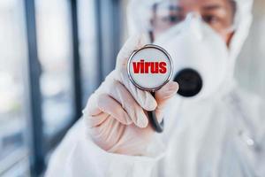 Krankenschwester in weißer Schutzkleidung, die drinnen steht, mit Stethoskop mit Virusbrief darauf foto