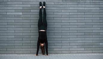 junges sportliches mädchen in schwarzer sportbekleidung, das im freien in der nähe der grauen wand harte handstandübungen macht foto