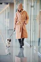 Frau in warmer Kleidung, die mit ihrem kleinen Pug-Hund drinnen in der Halle spazieren geht foto
