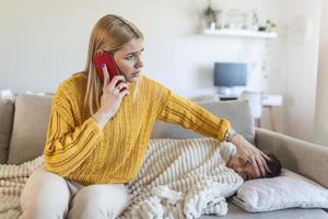 besorgte mutter, die die temperatur ihres kleinen sohnes misst, der mit fieber im bett liegt. junge mutter überprüft die temperatur ihres jungen mit thermometer auf einer couch im wohnzimmer und ruft arzt an. foto