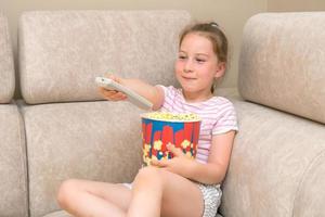 Kleines Mädchen sitzt mit einem großen Glas Popcorn auf der Couch und wechselt mit einem Lächeln die Fernsehkanäle auf der Fernbedienung foto