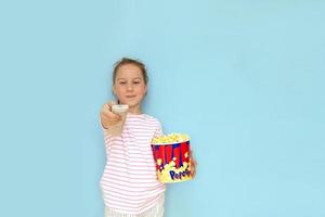 Kleines Mädchen hält ein großes Glas Popcorn in der einen Hand, während es mit der anderen die Fernbedienung des Fernsehers hält foto