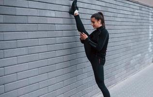 junges sportliches mädchen mit telefon in schwarzer sportbekleidung, das mit grauer wand die beine im freien streckt foto