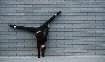 junges sportliches mädchen in schwarzer sportbekleidung, das im freien in der nähe der grauen wand harte handstandübungen macht foto