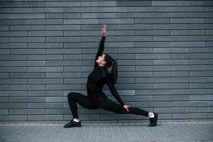 junges sportliches mädchen in schwarzer sportbekleidung im freien, das yoga-übungen in der nähe der grauen wand macht foto