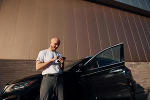 glatzköpfiger mann, der tagsüber neben seinem modernen schwarzen auto im freien steht, mit telefon in den händen foto