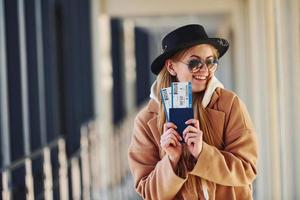 Junge Passagierin in warmer Kleidung, die Tickets in der Flughafenhalle zeigt foto