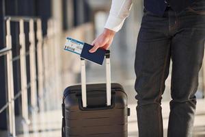 Nahaufnahme eines männlichen Passagiers in eleganter formeller Kleidung, der sich mit Gepäck und Tickets in der Flughafenhalle befindet foto