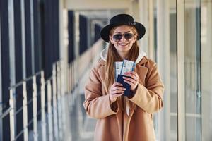 Junge Passagierin in warmer Kleidung, die Tickets in der Flughafenhalle zeigt foto