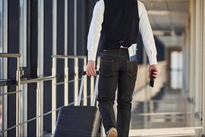 Rückansicht des männlichen Passagiers in eleganter formeller Kleidung befindet sich in der Flughafenhalle mit Tickets in der Tasche foto