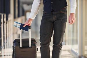 Nahaufnahme eines männlichen Passagiers in eleganter formeller Kleidung, der sich mit Gepäck und Tickets in der Flughafenhalle befindet foto