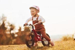 glückliches kleines Mädchen mit Schutzhut, das an einem sonnigen Tag in der Nähe des Waldes mit dem Fahrrad im Freien fährt foto