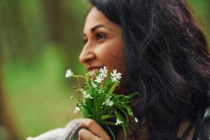 Fröhliche Brünette, die Blumen in den Händen hält, während sie zur Frühlingszeit im Wald steht foto