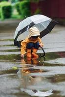 kind in gelbem wasserdichtem mantel, stiefeln und mit regenschirm, der nach dem regen im freien spielt foto