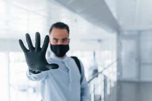 Stoppgeste von Hand zeigen. in Schutzmaske und Handschuhen. junger gutaussehender mann in formeller kleidung drinnen im büro tagsüber foto