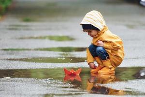 Kind in gelbem, wasserdichtem Mantel und Stiefeln, das nach dem Regen mit handgefertigtem Bootsspielzeug aus Papier im Freien spielt foto