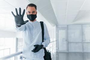 Stoppgeste von Hand zeigen. in Schutzmaske und Handschuhen. junger gutaussehender mann in formeller kleidung drinnen im büro tagsüber foto