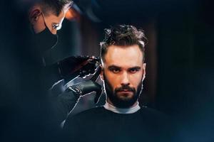 Vorderansicht des jungen bärtigen Mannes, der im Friseursalon von einem Mann in schwarzer Schutzmaske sitzt und sich die Haare schneiden lässt foto