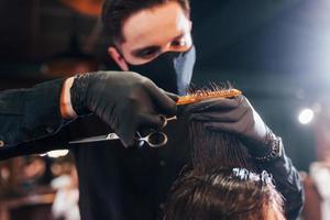 junger bärtiger mann, der im friseursalon sitzt und sich von einem kerl in schwarzer schutzmaske die haare schneiden lässt foto