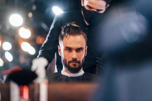 Vorderansicht des jungen bärtigen Mannes, der im Friseursalon von einem Mann in schwarzer Schutzmaske sitzt und sich die Haare schneiden lässt foto