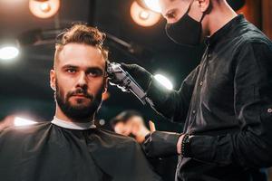 Vorderansicht des jungen bärtigen Mannes, der im Friseursalon von einem Mann in schwarzer Schutzmaske sitzt und sich die Haare schneiden lässt foto