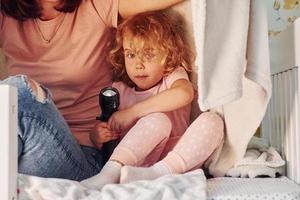 Sitzen unter der Decke mit Taschenlampe. junge mutter mit ihrer kleinen tochter in lässiger kleidung zusammen zuhause zu hause foto