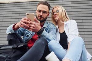 mit Telefon. junger stilvoller mann mit frau in lässiger kleidung, die zusammen im freien sitzt. Vorstellung von Freundschaft oder Beziehungen foto