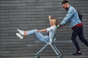 Spaß haben und Einkaufswagen fahren. junger stilvoller mann mit frau in lässiger kleidung im freien zusammen. Vorstellung von Freundschaft oder Beziehungen foto