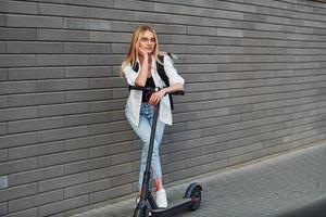 Schöne Blondine in lässiger Kleidung, die tagsüber bei sonnigem Wetter im Freien mit Elektroroller fährt foto