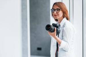 Frau in formeller Kleidung und mit Kamera in der Hand, die in einem leeren Raum steht foto