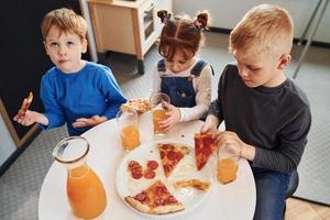 Drei Kinder sitzen drinnen am Tisch und essen gemeinsam Pizza mit Orangensaft foto