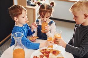 Drei Kinder sitzen drinnen am Tisch und essen gemeinsam Pizza mit Orangensaft foto