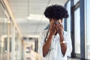 traurige und depressive junge afroamerikanische ärztin in weißer uniform, die im korridor steht foto