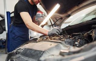 verwendet Beleuchtungsgeräte. Mann in Arbeitsuniform repariert weißes Auto drinnen. Konzept des Autoservices foto