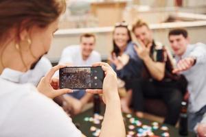 Mädchen macht Foto, wenn Leute Kartenspiel spielen. eine gruppe junger leute in lässiger kleidung feiert tagsüber zusammen eine party auf dem dach foto