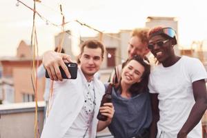 zusammenstehen und selfie machen. eine gruppe junger leute in lässiger kleidung feiert tagsüber zusammen eine party auf dem dach foto