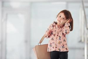 süßes kleines Mädchen mit Tasche drinnen im Büro oder am Flughafen, das per Telefon spricht foto