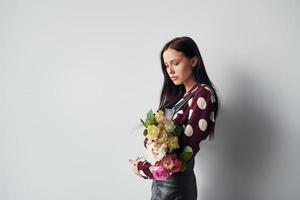 schöne junge Brünette im schwarzen Rock, die drinnen vor weißem Hintergrund steht, mit Blumenstrauß in den Händen foto