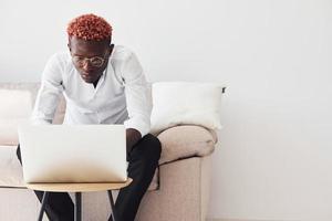 junger afroamerikanischer mann in formeller kleidung, der drinnen mit laptop in den händen sitzt foto