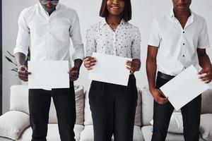 gruppe von afroamerikanern in formeller kleidung, die zusammen in der nähe des sofas drinnen im büro stehen und leere papiere in den händen halten foto