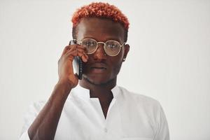 Junger afroamerikanischer Mann in weißer formeller Kleidung, der drinnen an der Wand telefoniert foto