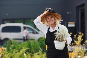 Topf in den Händen halten. Seniorin ist tagsüber im Minigarten. Gebäudehülle dahinter foto