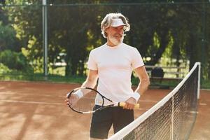 Älterer, moderner, stilvoller Mann mit Schläger im Freien auf dem Tennisplatz tagsüber foto